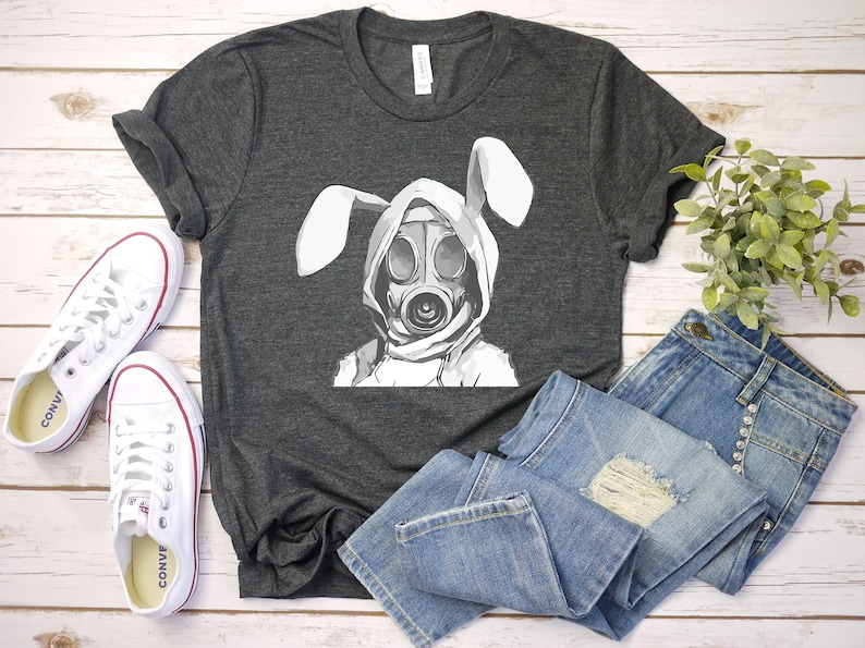 K&ouml;nnte beinhalten: Ein dunkelgraues T-Shirt mit einem wei&szlig;en Aufdruck eines Kaninchens, das eine Gasmaske tr&auml;gt. Das Kaninchen hat lange, schlaffe Ohren. Das T-Shirt liegt auf einer Holzoberfl&auml;che mit einem Paar wei&szlig;en Turnschuhen und einer blauen Jeans.
