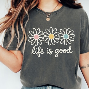 A woman wearing a gray t-shirt with a graphic design of three white flowers and the text "life is good" printed on it.