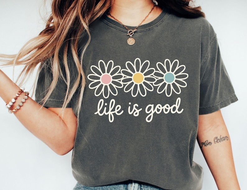 A woman wearing a gray t-shirt with a graphic design of three white flowers and the text "life is good" printed on it.