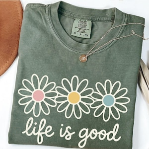 a green t-shirt with a flower design and the text "life is good" printed on it, along with a necklace and a hat.