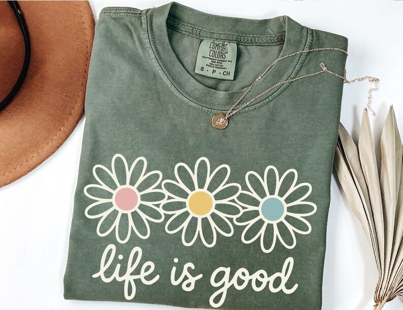 a green t-shirt with a flower design and the text "life is good" printed on it, along with a necklace and a hat.