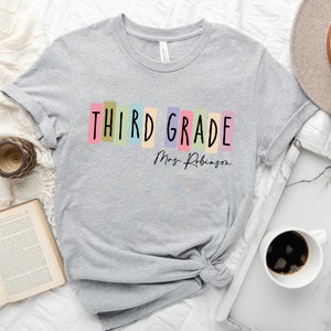 Op de afbeelding: Grijs T-shirt met de tekst "THIRD GRADE" in kleurrijke blokken en "Mrs. Robinson" in zwarte schrijfletters.