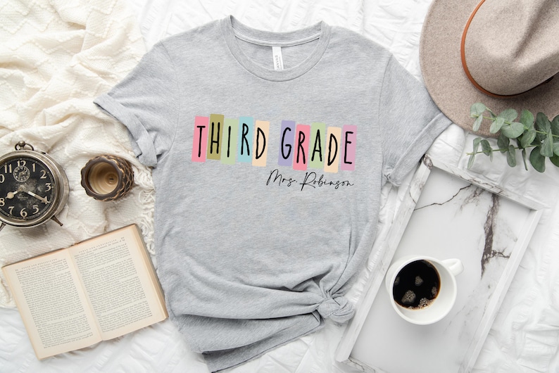 Op de afbeelding: Grijs T-shirt met de tekst "THIRD GRADE" in kleurrijke blokken en "Mrs. Robinson" in zwarte schrijfletters.