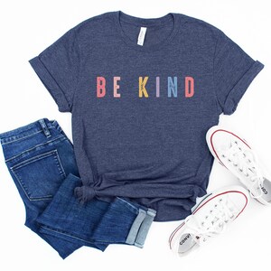 May include: A navy blue t-shirt with the words "BE KIND" printed in rainbow colors. The shirt is paired with blue jeans and white sneakers.