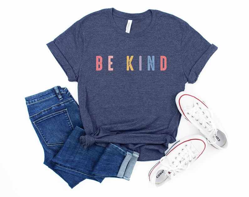 May include: A navy blue t-shirt with the words "BE KIND" printed in rainbow colors. The shirt is paired with blue jeans and white sneakers.