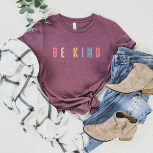 May include: A maroon t-shirt with the words "BE KIND" in rainbow colors. The shirt is styled with a white and black plaid scarf, blue jeans, and brown ankle boots.