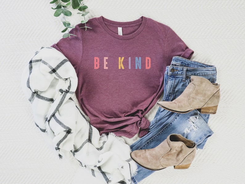 May include: A maroon t-shirt with the words "BE KIND" in rainbow colors. The shirt is styled with a white and black plaid scarf, blue jeans, and brown ankle boots.