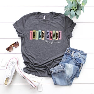 Op de afbeelding: Een donkergrijs T-shirt met de tekst "THIRD GRADE" in kleurrijke blokletters en "Mrs. Robinson" in witte tekst eronder. Het shirt ligt plat op een wit houten oppervlak met een zonnebril, witte sneakers en een blauwe jeans.