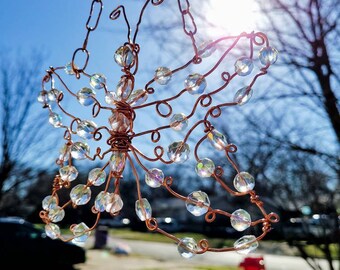 May include: A copper wire butterfly mobile with clear crystal beads hanging from a copper chain. The mobile is outdoors with a blurred background of green grass and trees.