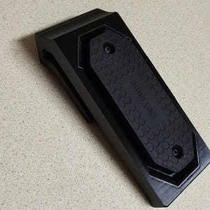 May include: Black rectangular device with a textured, rubberized top and the word "DeYoung" printed on it. The device has a beveled edge and two small holes. The item is on a light-colored surface.