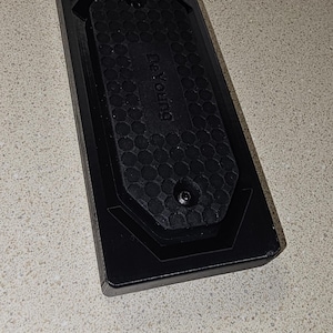 May include: A black rectangular object with a textured surface and the word "ReYoung" printed on it. The object has a hexagonal pattern in the center and two screws. The item is likely a part of a larger product.