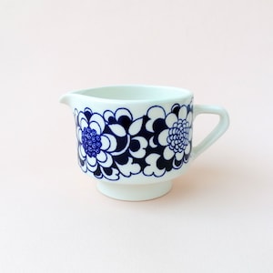 Puede incluir: Una jarra de crema de cerámica blanca con un diseño floral azul y blanco. La jarra tiene un asa y una base redonda.