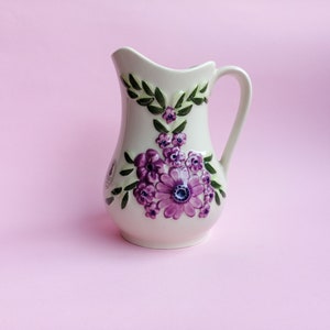 Puede incluir: Una jarra de cerámica blanca con un diseño floral. La jarra tiene un asa y está decorada con flores moradas y hojas verdes.