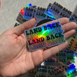 Puede incluir: Una pegatina hologr&aacute;fica con el texto "LAND BACK" repetido cuatro veces en un esquema de colores arco&iacute;ris. La pegatina se sostiene en una mano.