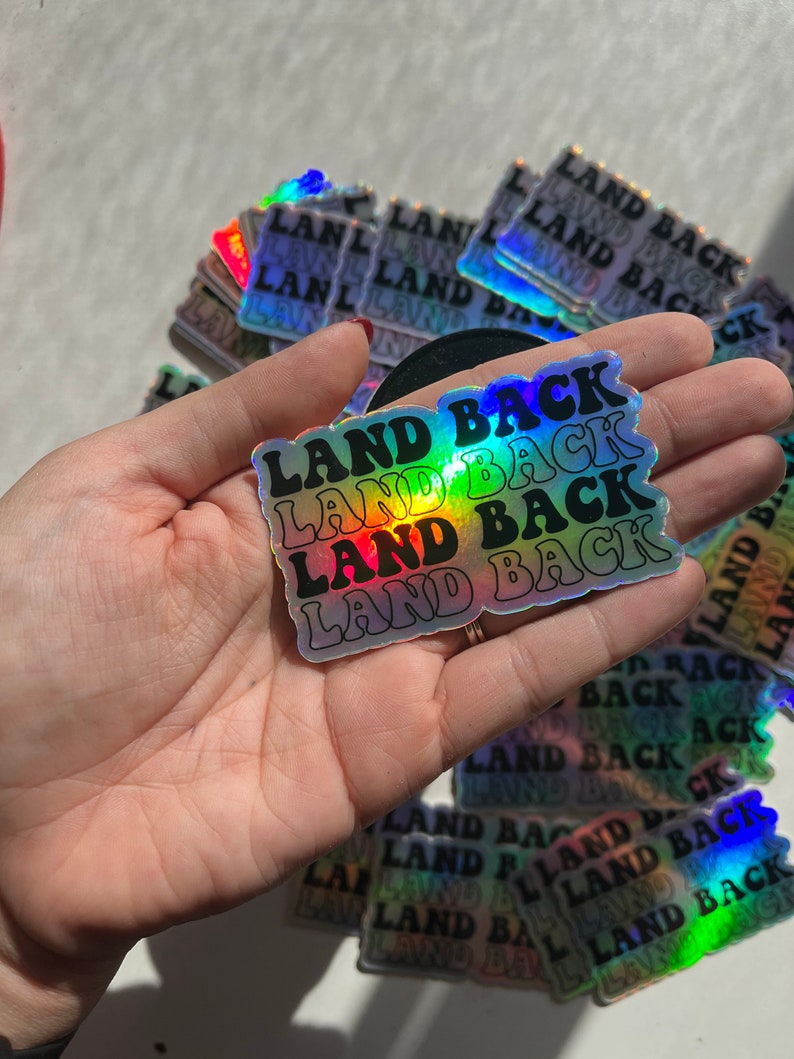 Puede incluir: Una pegatina hologr&aacute;fica con el texto "LAND BACK" repetido cuatro veces en un esquema de colores arco&iacute;ris. La pegatina se sostiene en una mano.