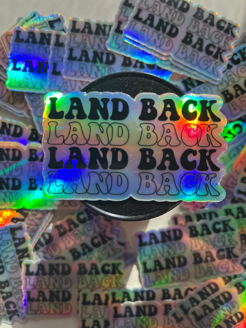 Puede incluir: Primer plano de varias pegatinas iridiscentes con el texto "Land Back" repetido en un dise&ntilde;o hologr&aacute;fico arco&iacute;ris.