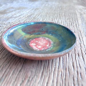 May include: A small, handmade ceramic bowl with a vibrant, colourful interior. The bowl features a red centre, surrounded by rings of yellow, green, blue, and black. The rim is a reddish-brown colour. The bowl is set on a wooden surface.