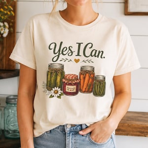 Peut inclure: Un t-shirt à manches courtes de couleur crème avec le texte Yes I Can en écriture vert foncé. Sous le texte se trouvent des illustrations de quatre bocaux en verre remplis de cornichons, de confiture rouge, de carottes et de haricots verts.