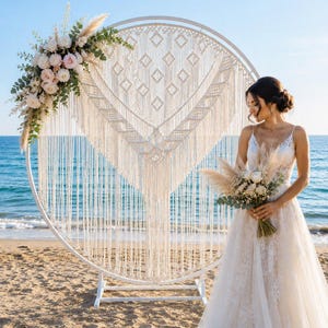 May include: A large circular white metal frame featuring an intricate off-white macrame tapestry with geometric patterns. The frame is adorned with a floral arrangement of white and pink roses, pampas grass, and green eucalyptus leaves.
