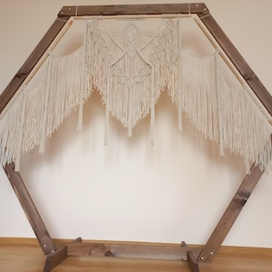 Könnte beinhalten: Ein sechseckiger Holzbogen mit einer Makramee-Wandbehang. Der Wandbehang ist aus weißem Baumwollgarn gefertigt und hat ein geometrisches Muster.