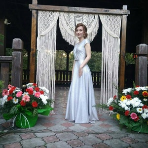 以下が含まれることがあります： 銀色のスパンコールのドレスを着た女性が、マクラメの結婚式のアーチの前に立っています。彼女は微笑んでカメラを見ています。彼女の両側には花束があります。