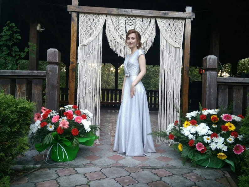 以下が含まれることがあります： 銀色のスパンコールのドレスを着た女性が、マクラメの結婚式のアーチの前に立っています。彼女は微笑んでカメラを見ています。彼女の両側には花束があります。