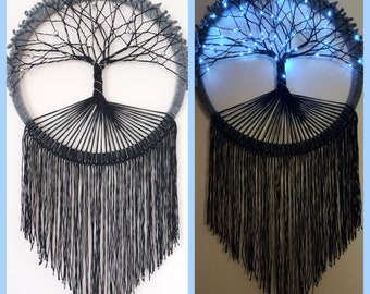 May include: A gray and black macrame wall hanging with a tree of life design. The tree is decorated with small white beads and the hanging has black fringe. The second image shows the same hanging with blue lights illuminating the tree.