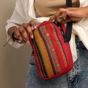 May include: A woman is holding a red and yellow striped crossbody bag with a black strap. The bag has a geometric pattern and is made of fabric. The bag is worn over the shoulder and the woman is looking down at it.