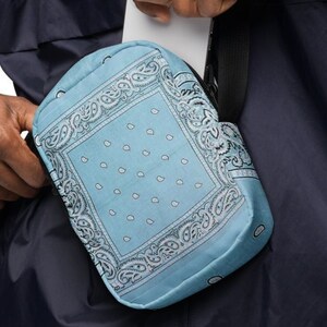 May include: A blue and white bandana print crossbody bag with a black adjustable strap. The bag is worn by a person wearing a dark blue jacket.