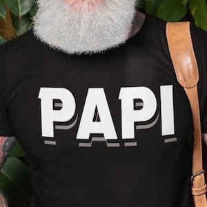 May include: A black t-shirt with the word "PAPI" printed in white with a shadow effect. The shirt is being worn by a person with a white beard and tattoos.