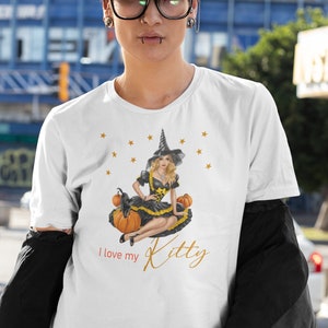 May include: White t-shirt with a graphic of a witch in a black dress and hat sitting on pumpkins with a black cat. The text "I love my Kitty" is printed on the shirt.