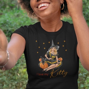 May include: Black t-shirt with a graphic of a witch sitting on pumpkins with a black cat. The text "I love my Kitty" is printed on the shirt.
