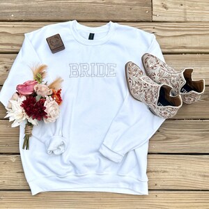 Könnte beinhalten: Ein weißes Sweatshirt mit dem Wort "BRIDE" in gestickten Großbuchstaben. Ein kleiner Blumenstrauß und ein Paar cremefarbene Cowboy-Stiefel mit detaillierten floralen Mustern sind ebenfalls zu sehen.