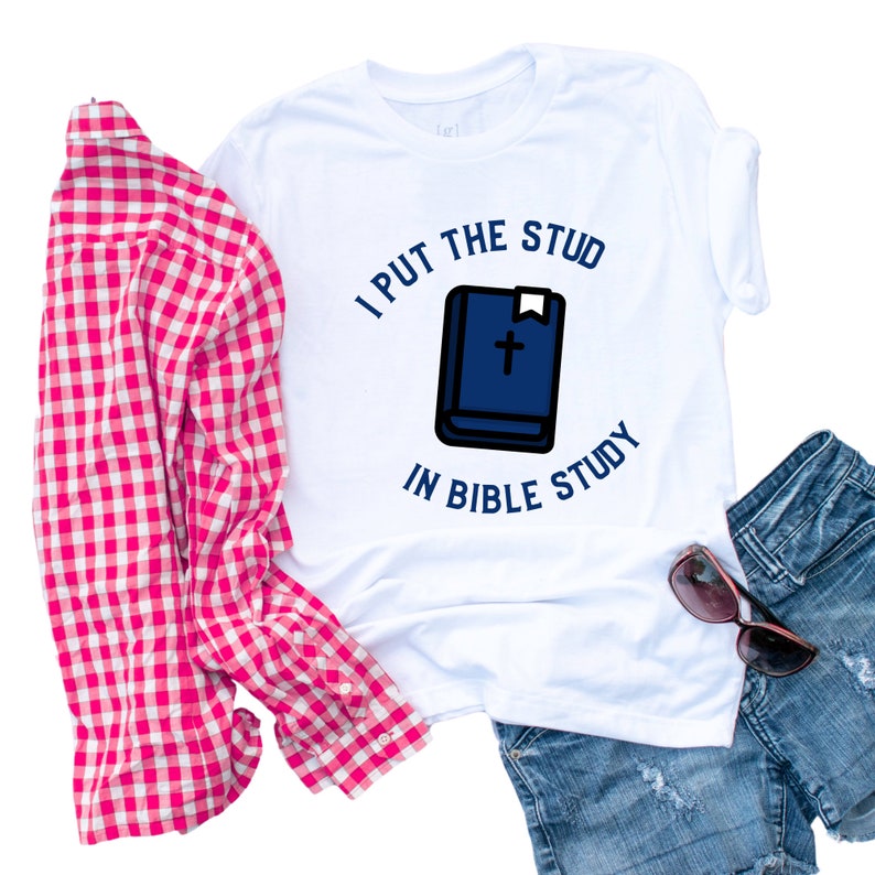 Pode incluir: Camiseta branca com um gr&aacute;fico azul e branco que diz "I put the stud in bible study". O gr&aacute;fico inclui uma b&iacute;blia azul com uma cruz branca e um marcador.