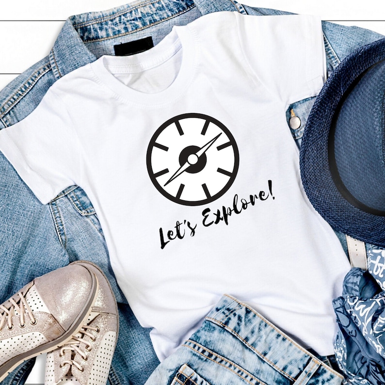 May include: White t-shirt with a black compass design and the text "Let's Explore!" printed on it. The shirt is laid out on a denim background with a hat and shoes.