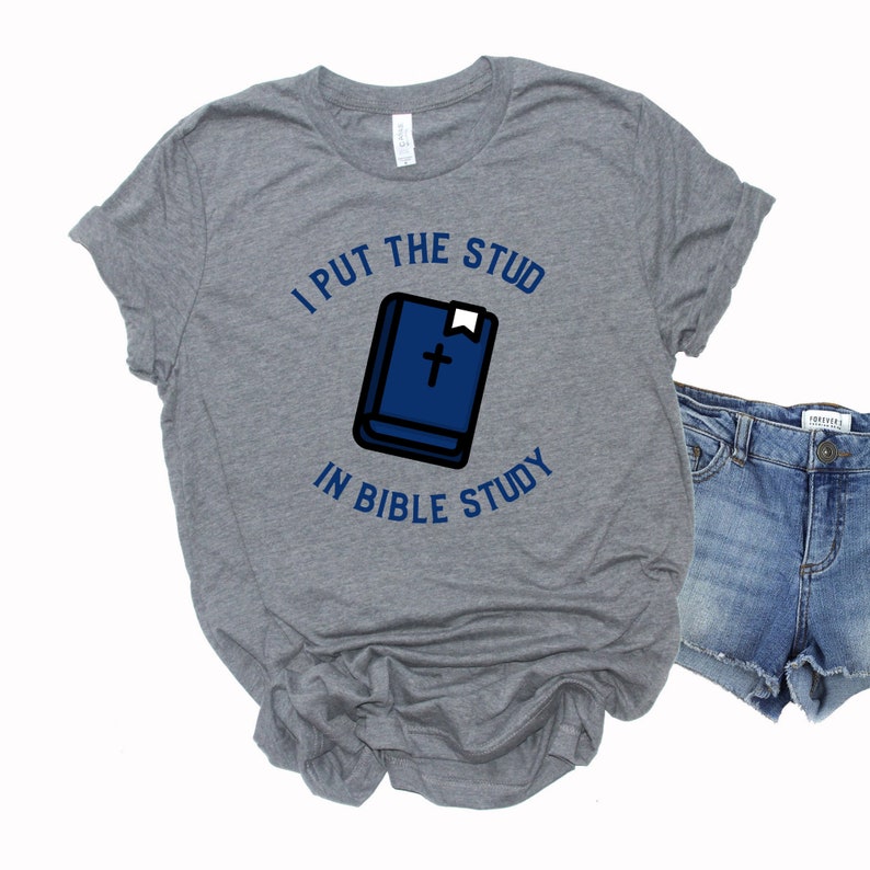 Pode incluir: Uma camiseta cinza com o texto "I put the stud in bible study" e uma ilustra&ccedil;&atilde;o azul de uma b&iacute;blia com uma cruz na capa.