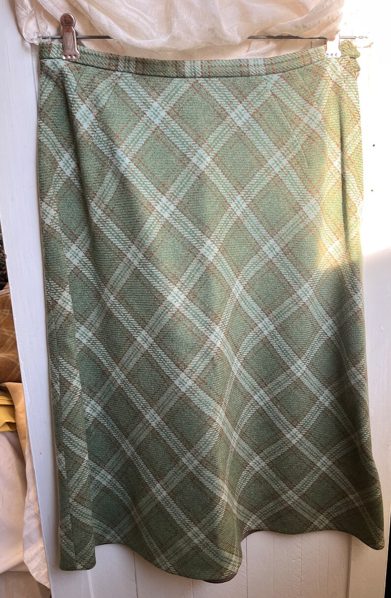 Puede incluir: Una falda de cuadros verdes con un patr&oacute;n de cuadros blancos y marrones. La falda es de largo hasta la rodilla y tiene una silueta recta.