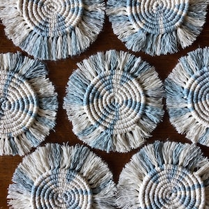 May include: Set of six blue and white crocheted coasters with fringe. The coasters are circular and have a woven pattern.