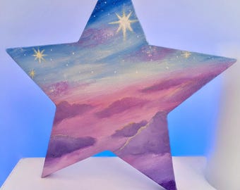 May include: A star-shaped painting with a celestial theme. The artwork features a gradient of colors, including blue, purple, and pink, depicting a starry sky and clouds. Several white stars are scattered across the canvas, adding to the dreamy aesthetic.
