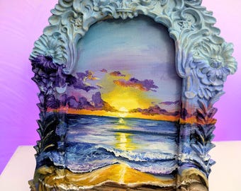 May include: Ornate, blue-gray framed artwork depicting a sunset over the ocean. The painting features a vibrant sky with yellow, orange, and purple hues, reflecting on the water. The frame has detailed scrollwork and a brown base.