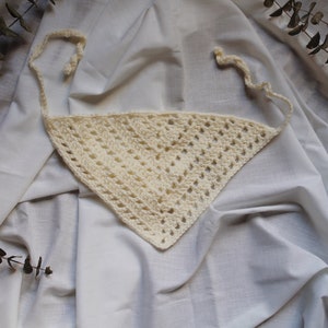 May include: A white crocheted triangle scarf with a fringe on the edges. The scarf is lying on a white fabric background.