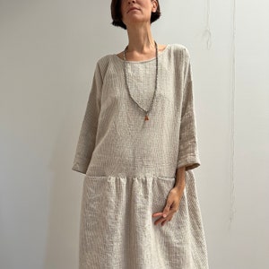 May include: A beige, textured, long-sleeve dress with a scoop neck and pockets. The dress has a gathered waist and falls to the mid-calf. A long beaded necklace with a pendant is worn over the dress.