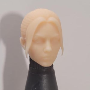 May include: A pale-toned, sculpted head with long, swept-back hair and a neutral expression. The head is attached to a black, cylindrical base. The hair is styled with a side part and long bangs. The overall aesthetic is smooth and detailed.