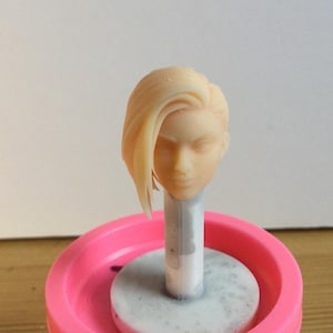 May include: A light-colored, unpainted resin miniature head sculpt featuring a short, asymmetrical hairstyle with a side-swept fringe. The head is mounted on a vertical white cylindrical post, which is secured within a bright pink circular base.