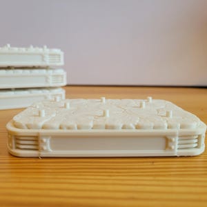 May include: A rectangular off-white display base featuring a textured, rocky surface with several small vertical pegs for mounting. The sides of the base have a layered, ribbed design with a smooth central panel. Multiple identical bases are stacked behind.