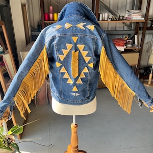 Puede incluir: Una chaqueta vaquera azul con mangas con flecos amarillos y diseños geométricos en la espalda. La chaqueta se exhibe en un maniquí de madera. La espalda presenta formas geométricas amarillas y blancas.