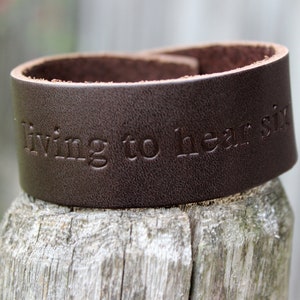 Puede incluir: Un brazalete de cuero marrón con las palabras "living to hear six" grabadas en la parte delantera.