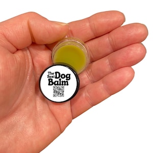 May include: A small, clear jar filled with a light green balm, held in a hand. The jar's black lid features the text "The Best Dog Balm" and a QR code. The balm appears to be a skincare product for dogs.