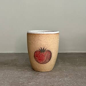 May include: A beige ceramic mug with a speckled glaze and a white rim. The mug features a hand-drawn illustration of a red tomato.