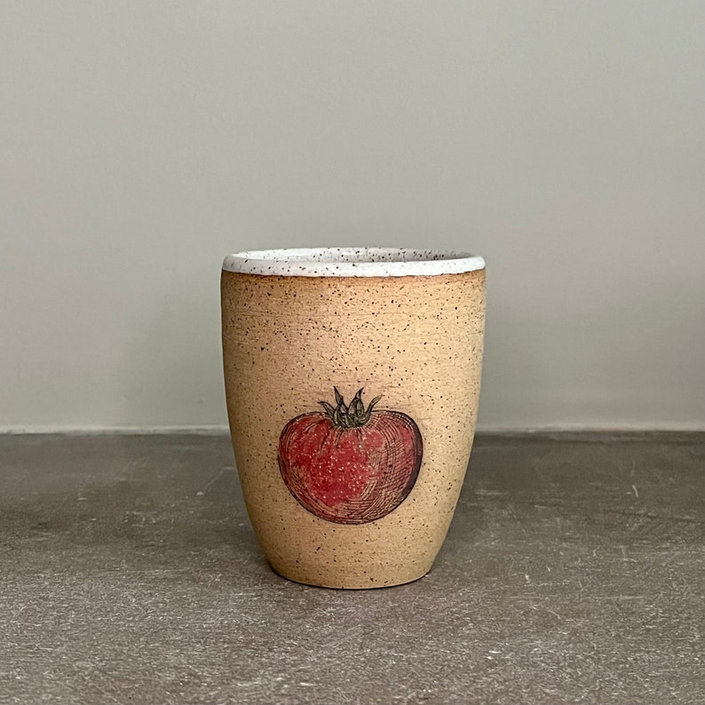 May include: A beige ceramic mug with a speckled glaze and a white rim. The mug features a hand-drawn illustration of a red tomato.